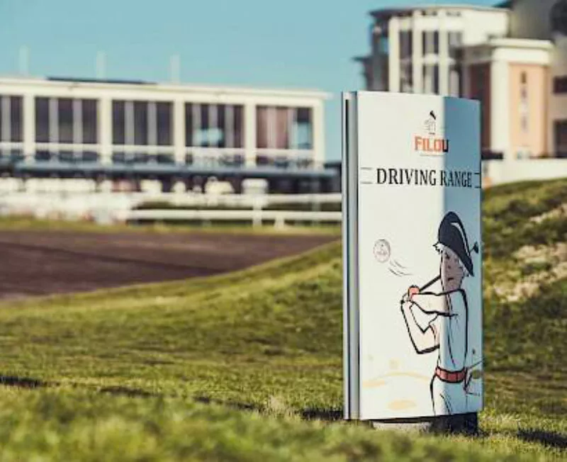 Bord met opschrift ‘Filou Driving Range’ op grasveld, met illustratie van Filou-mannetje in een witte golfoutfit die een golfclub zwaait; op de achtergrond een groot gebouw met ramen en balkons – onderdeel van Wellington Golf en Filou Golf beleving.