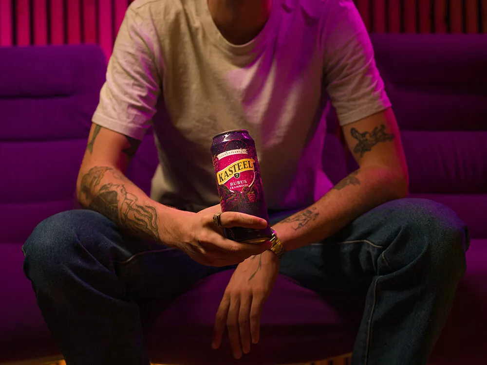 Een man met tattoos houdt een blik Kasteel Rubus Framboise vast terwijl hij zit op een paarse zetel. Je ziet zijn hoofd niet, zodat het blik duidelijk in beeld is. Kasteel Rubus Framboise is een sterk fruitbier gebrouwen met echte frambozen. 