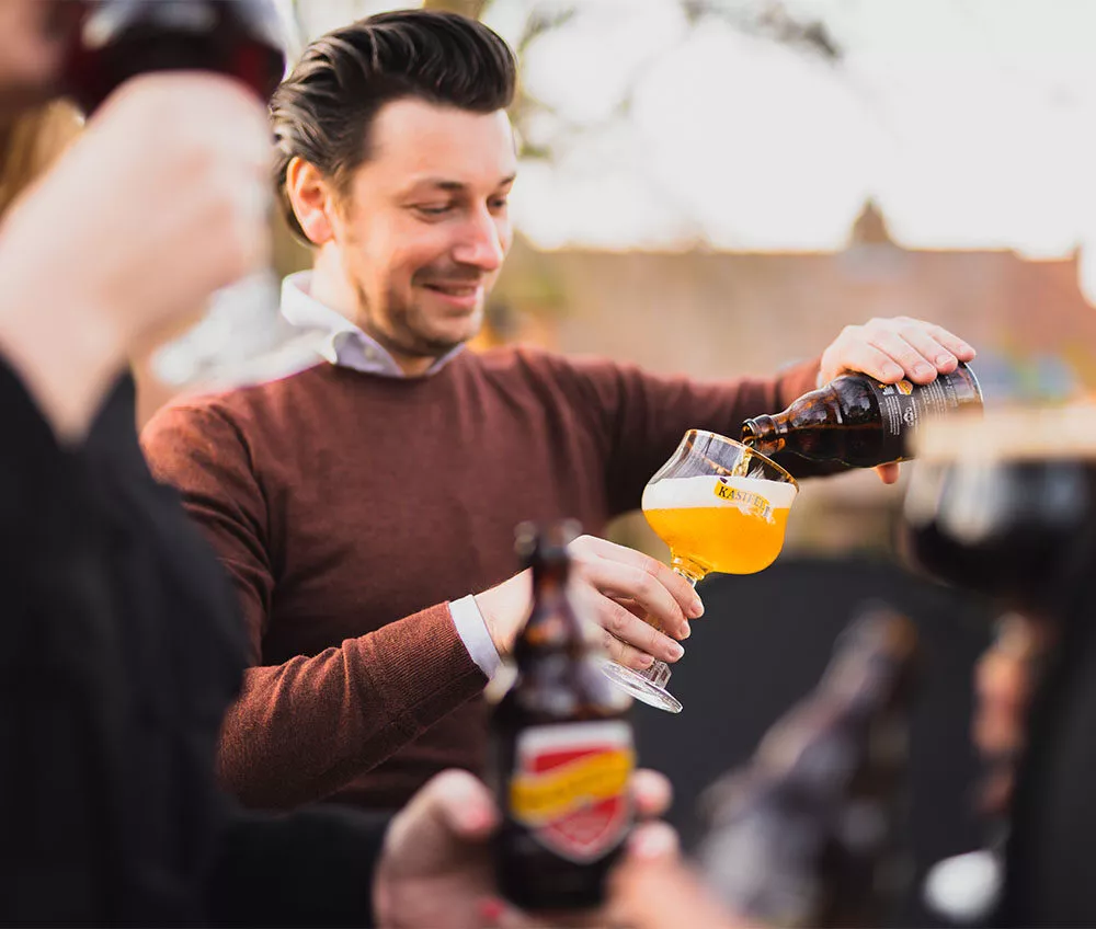Man schenkt een 33 cl fles Kasteel Tripel uit waardoor het bierglas van Kasteel zich vult met een helder honingkleurig bier met een fijne witte schuimkraag. Op de voorgrond zie je een wazige 33 cl fles Kasteel Rouge in beeld, die wordt vastgehouden door een persoon die met de andere hand nipt van een gevuld glas Kasteel Rouge.