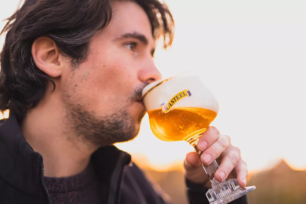 Een man met baard en snor nipt van een glas Kasteel Tripel, dat is gevuld met een honingkleurig bier met een dikke witte schuimkraag. De avondzon werpt een mooi licht op het bier.