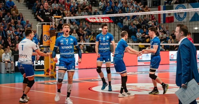 Spelers van Knack Volley Roeselare in blauwe shirts , actief op het volleybalveld tijdens een indoorwedstrijd; libero in wit shirt en publiek op de achtergrond – Filou volleybal sponsoring.