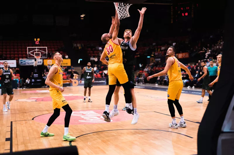 Speler van Basketbalclub Filou Oostende in geel shirt met rugnummer 30 probeert te scoren tijdens een wedstrijd, verdedigd door een tegenstander; andere spelers en publiek zichtbaar op de achtergrond – branding van Filou bier gebrouwen door Kasteel Brouwerij Vanhonsebrouck.