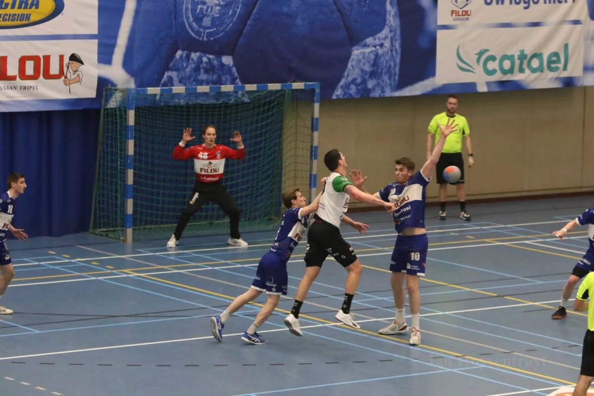 Twee spelers van Handbalclub Filou Izegem (HBCI) in blauw voorkomen dat de tegenstander scoort; doelman in rood-zwart staat klaar voor het doel, met banner van Filou bier op de achtergrond.