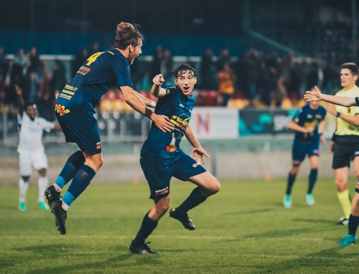Voetbalwedstrijd met spelers van KV Diksmuide Oostende (KVDO) in blauwe shirts, waarbij één speler springt en een andere loopt; andere spelers en toeschouwers zichtbaar op de achtergrond – Filou voetbalbeleving.