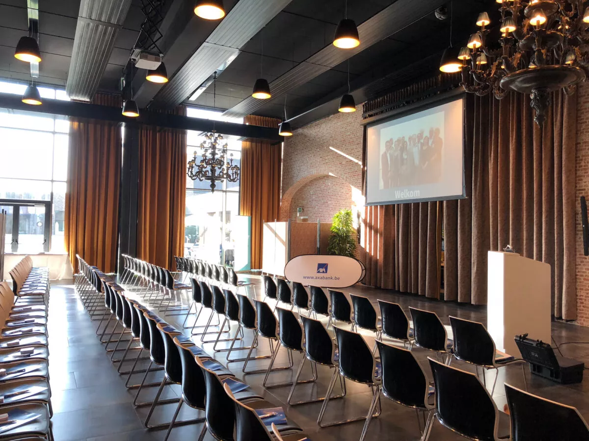 Balzaal van het Bierkasteel in Izegem ingericht voor een bedrijfsevent met rijen zwarte stoelen, podium, groot scherm en natuurlijke lichtinval via hoge ramen.