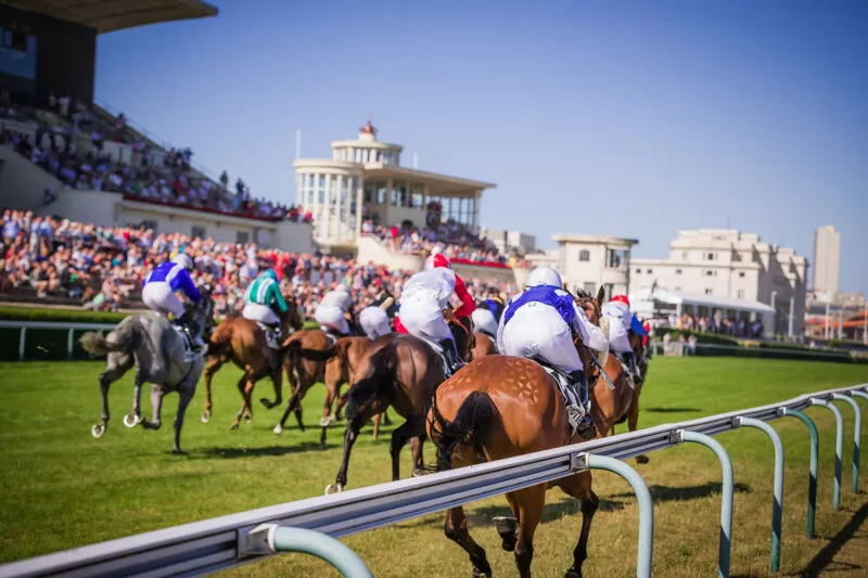 Paardenrennen tijdens Oostende Koerse met jockeys in kleurrijke tenues op galopperende paarden, racetrack met witte reling, volle tribunes met toeschouwers en gebouwen op de achtergrond – zonnige dag in Oostende, sfeer van Filou paardrijden.