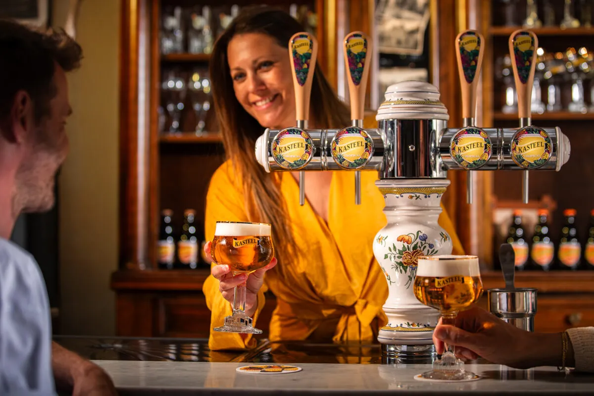 Barvrouw serveert Kasteel Tropical, een Kasteelbier gebrouwen met mango, passievrucht, perzik en ananas, aan klanten in een sfeervolle bar met tapinstallatie en flessenrek op de achtergrond.