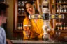 Barvrouw serveert Kasteel Tropical, een Kasteelbier gebrouwen met mango, passievrucht, perzik en ananas, aan klanten in een sfeervolle bar met tapinstallatie en flessenrek op de achtergrond.