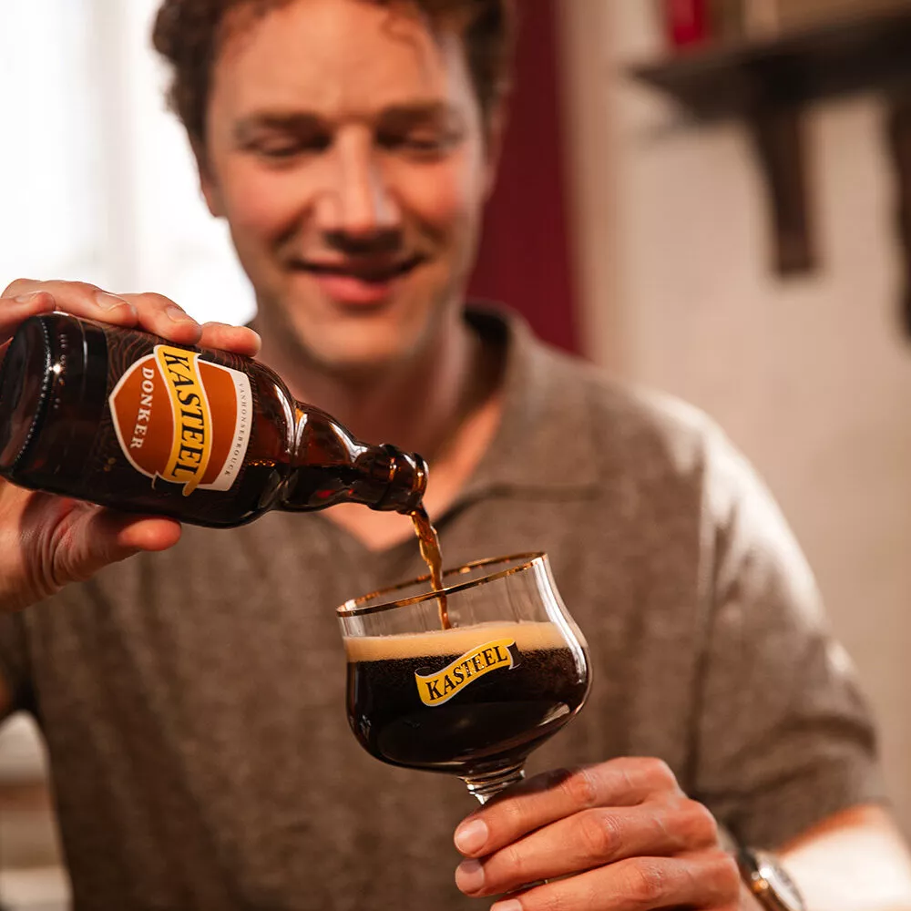 Een man schenkt een 33 cl fles Kasteel Donker uit in een bierglas van Kasteel, waarop je het logo - een wimpel met de letters 'KASTEEL' in - van het Belgische biermerk ziet. Het glas vult zich met een 