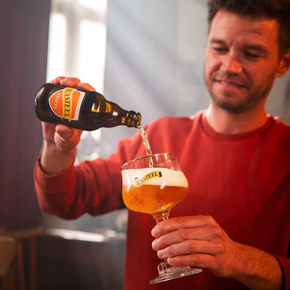 Een man schenkt Kasteel Tripel uit in een glas van Kasteel, een biermerk dat wordt gebrouwen door Kasteel Brouwerij Vanhonsebrouck. Het glas vult zich met een goudblond  een fles in een glas met schuimkraag – goudblond Belgisch abdijbier met karakter
